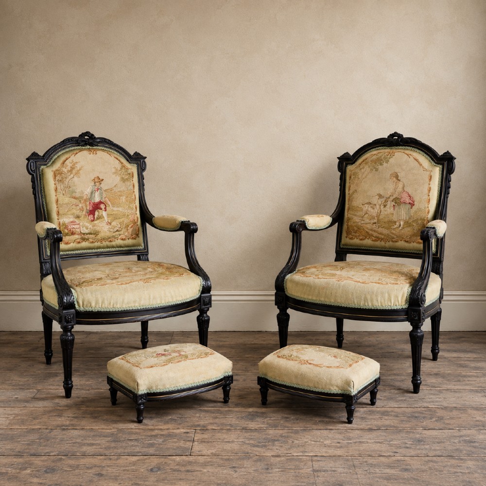 A pair of ebonised beech Louis XVI Revival salon armchairs featuring carved frames, fluted legs, padded armrests, and tapestry upholstery, accompanied by matching footstools.