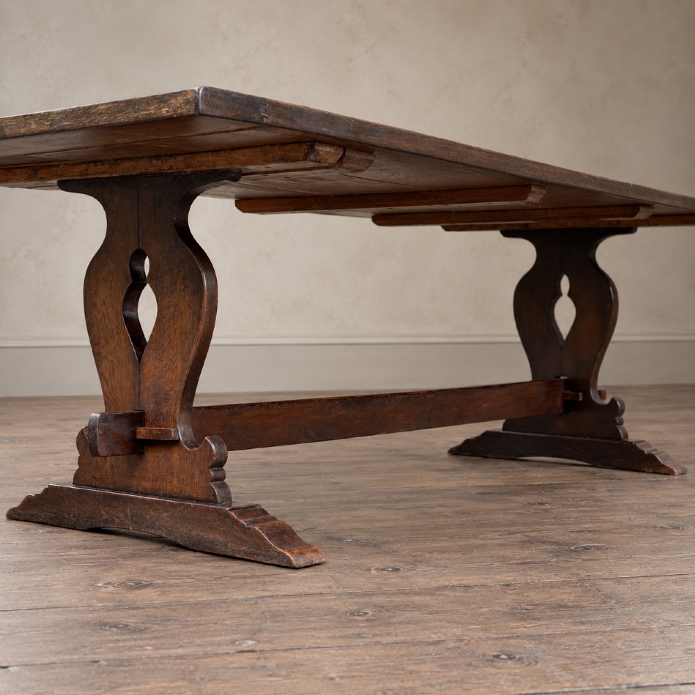 An early twentieth-century dark oak refectory dining table featuring a plank top, pierced trestle end supports, and a central connecting stretcher. View XII
