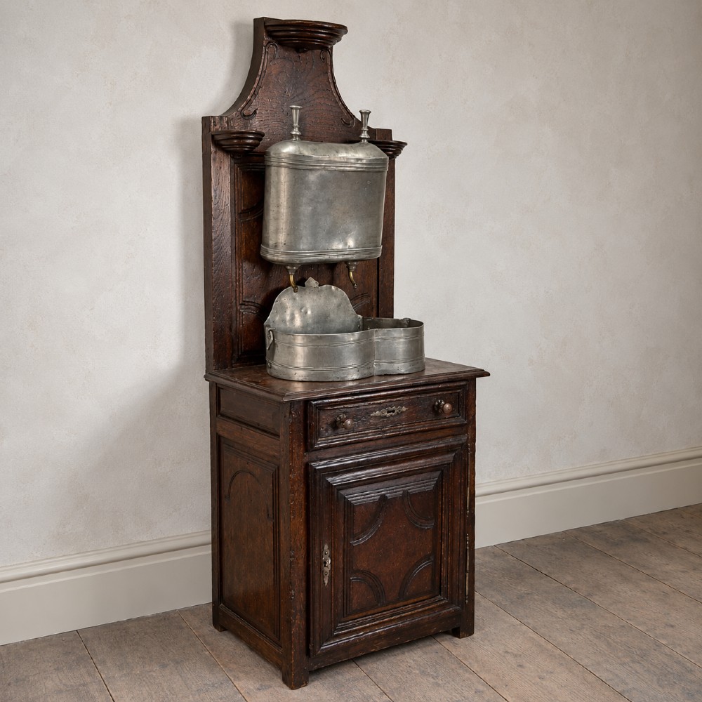 An 18th-century dark chestnut wood lavabo cabinet featuring a carved backboard mounted with a pewter cistern above a matching pewter basin. View V