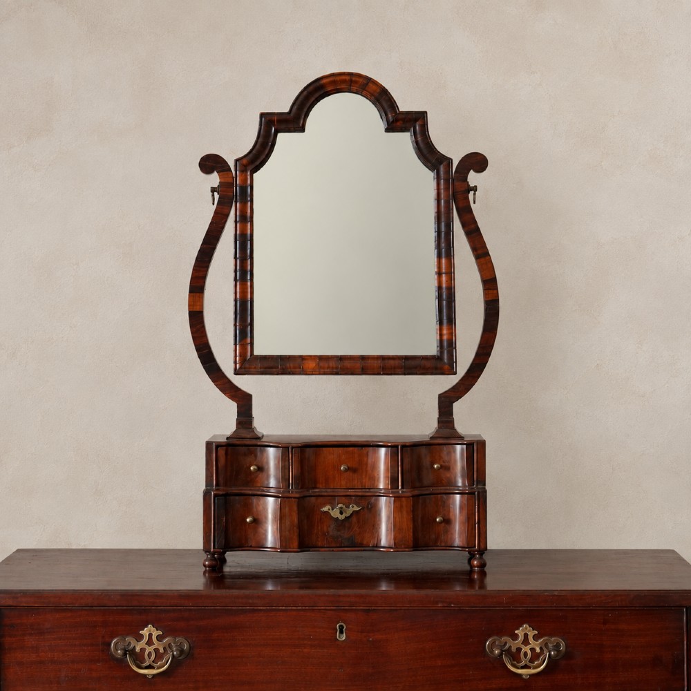 An antique Queen Anne walnut and laburnum toilet mirror featuring an arched cross-banded frame above a serpentine base with miniature drawers.