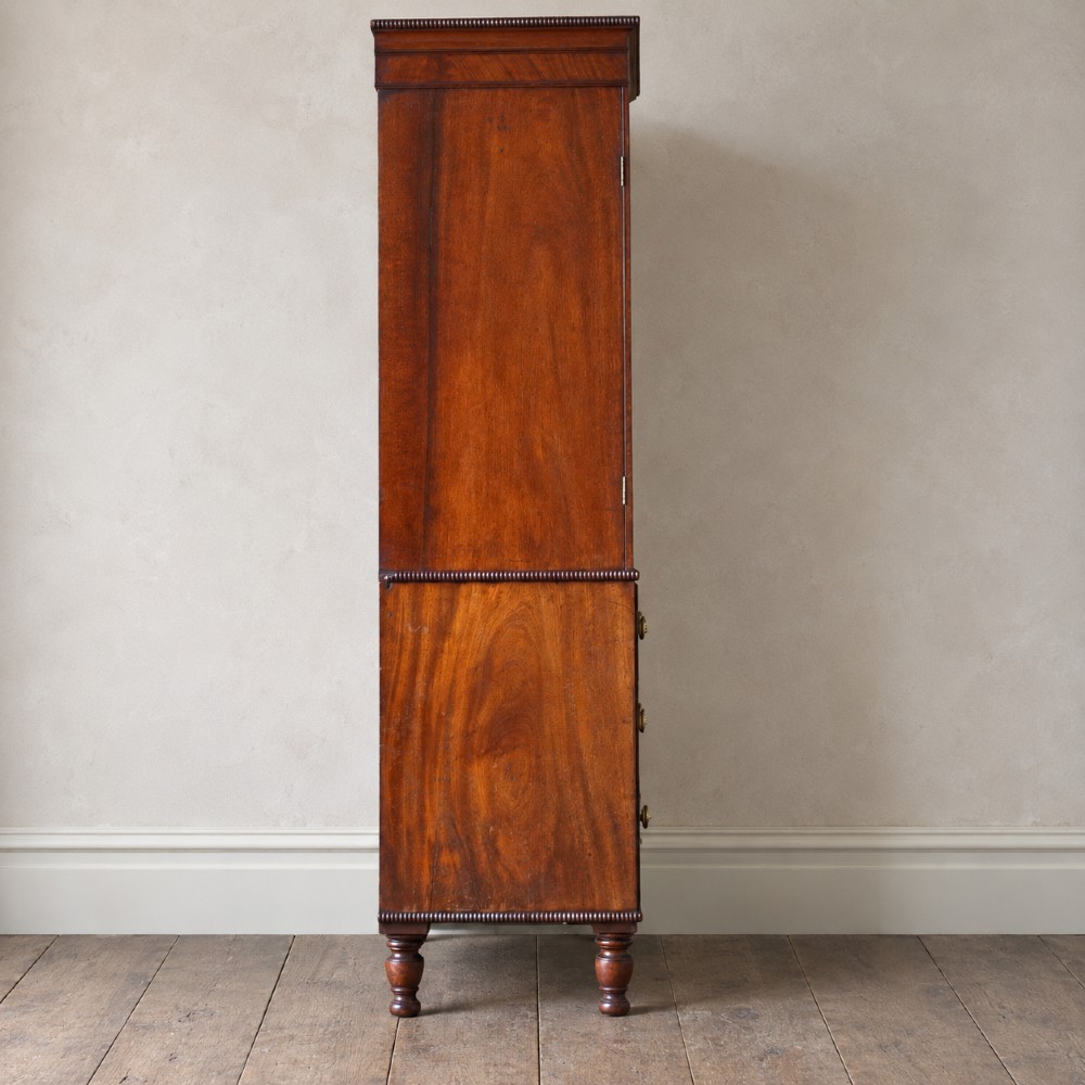 A Regency period flame mahogany linen press featuring a two-door upper cupboard above four drawers resting on turned legs. View VI