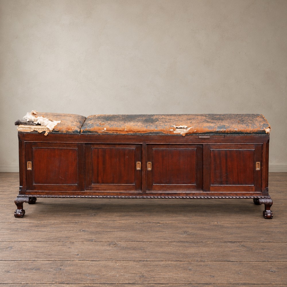 A long antique dark mahogany bench with four panelled cupboard doors, carved ball and claw feet, and a worn leather cushion top with a raised headrest.