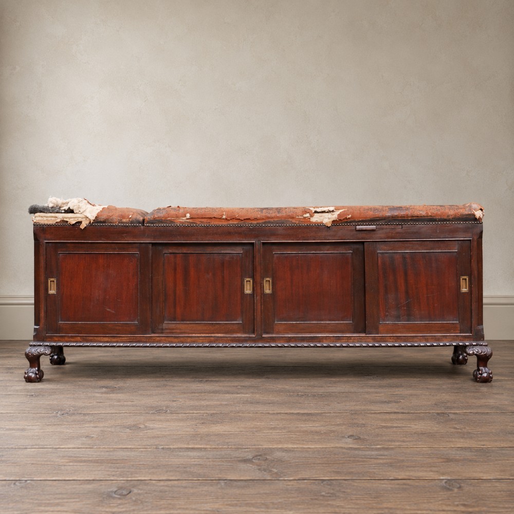 A long antique dark mahogany bench with four panelled cupboard doors, carved ball and claw feet, and a worn leather cushion top with a raised headrest. View IV
