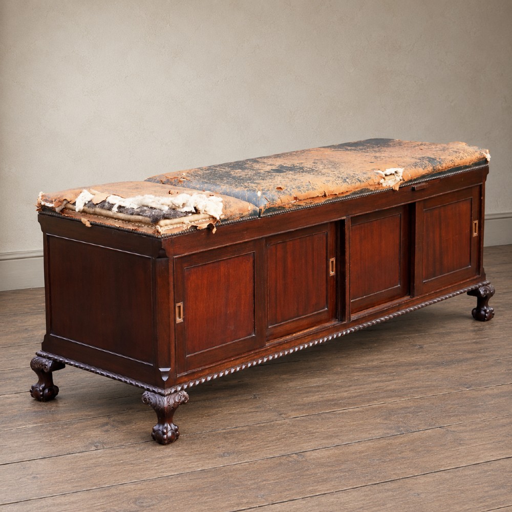 A long antique dark mahogany bench with four panelled cupboard doors, carved ball and claw feet, and a worn leather cushion top with a raised headrest. View V