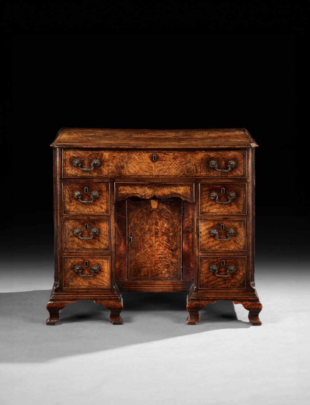 A George II fiddleback walnut kneehole desk featuring a central recessed cupboard, upper long drawer, six side drawers, and brass handles.