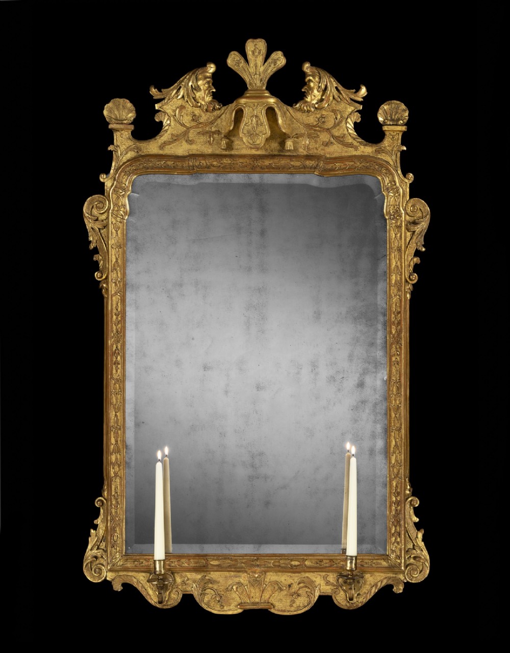 An 18th-century George I gilt gesso wall mirror featuring an ornate carved pediment, side scrolls, aged mirror plate, and two brass candle sconce nozzles.