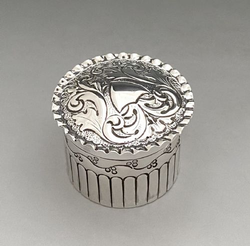 fig 1. A circular Victorian sterling silver pill box decorated with foliate scroll engraving, an unengraved shield cartouche, a crimped rim, and vertical fluting around the base.