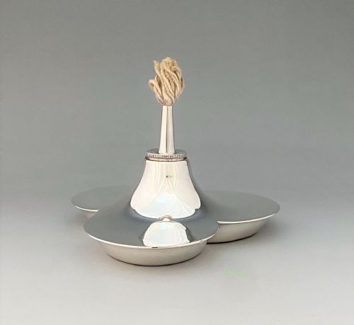 fig 1. A polished silver table lighter featuring a three-lobed trefoil base and a central upright nozzle holding a thick cotton wick.