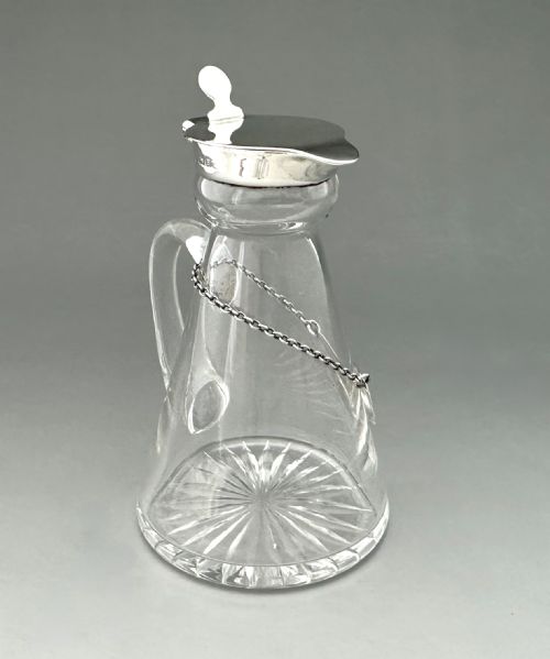 fig 1. A clear glass conical whisky tot with a star-cut base, a loop handle, a hinged sterling silver lid, and a silver label inscribed with Whisky.