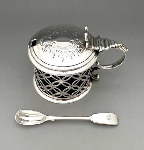 fig 1. A William IV sterling silver drum mustard pot featuring openwork geometric fretwork, a fitted blue glass liner, an engraved flat lid with a thumbpiece, a scroll handle, and a silver spoon.