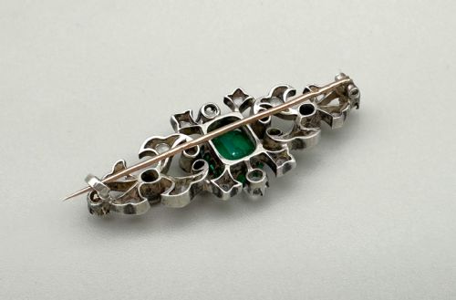 fig 1. An antique French elongated brooch crafted from silver-topped gold, featuring a central cushion-cut green emerald surrounded by pierced scrollwork set with bright stones.
