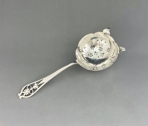 fig 1. A silver tea strainer featuring a pierced bowl with geometric drainage holes and a decoratively cut-out flat handle.