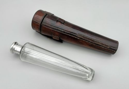 fig 1. A conical clear glass hunting flask with a silver cap positioned next to its fitted dark brown leather saddle holster.