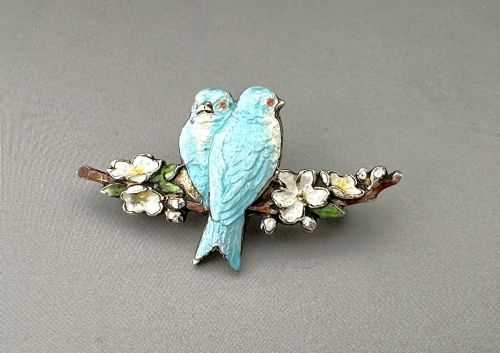 fig 1. A silver-gilt brooch depicting two light blue enamel birds perched on a flowering branch with white enamel blossoms.