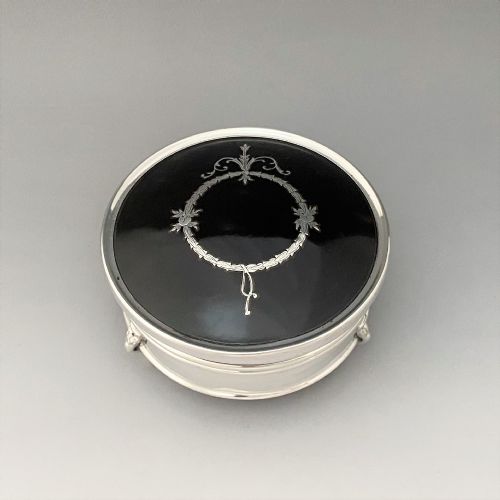 fig 1. A circular sterling silver trinket box resting on four cabriole legs featuring a dark tortoiseshell lid inlaid with a silver floral garland motif.