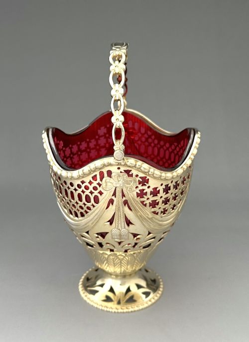 fig 1. A pierced silver-gilt sugar basket featuring drape and bow motifs with an overhead swing handle, housing a scalloped ruby red cranberry glass liner.