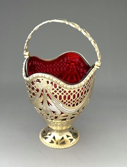 fig 2. A pierced silver-gilt sugar basket featuring drape and bow motifs with an overhead swing handle, housing a scalloped ruby red cranberry glass liner.