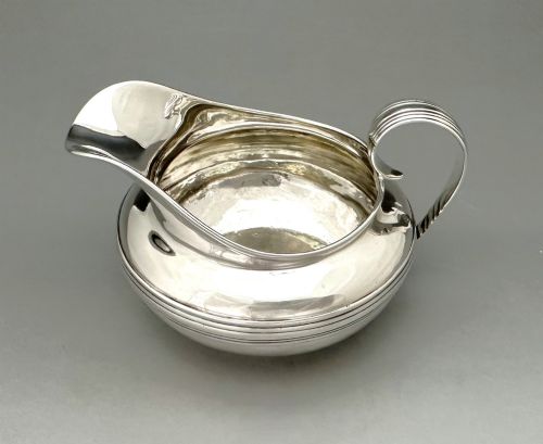 fig 1. A George IV sterling silver cream jug with a polished surface, horizontal reeded decoration on the waist, a wide pour spout, and a curved high-loop handle.