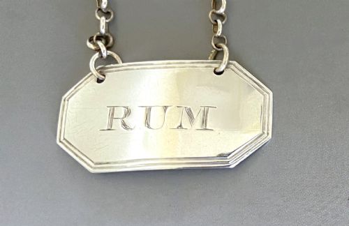 fig 1. An octagonal sterling silver decanter label engraved with the word RUM, featuring a reeded border and suspended by a thin silver chain.