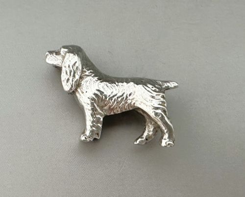 fig 1. A cast silver brooch finely detailed in the shape of a standing spaniel dog with textured fur.