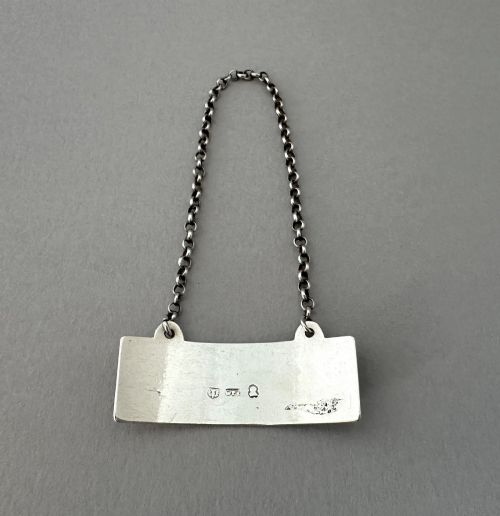 fig 1. A rectangular silver decanter label engraved with the word PORT and suspended by a thin silver link chain.