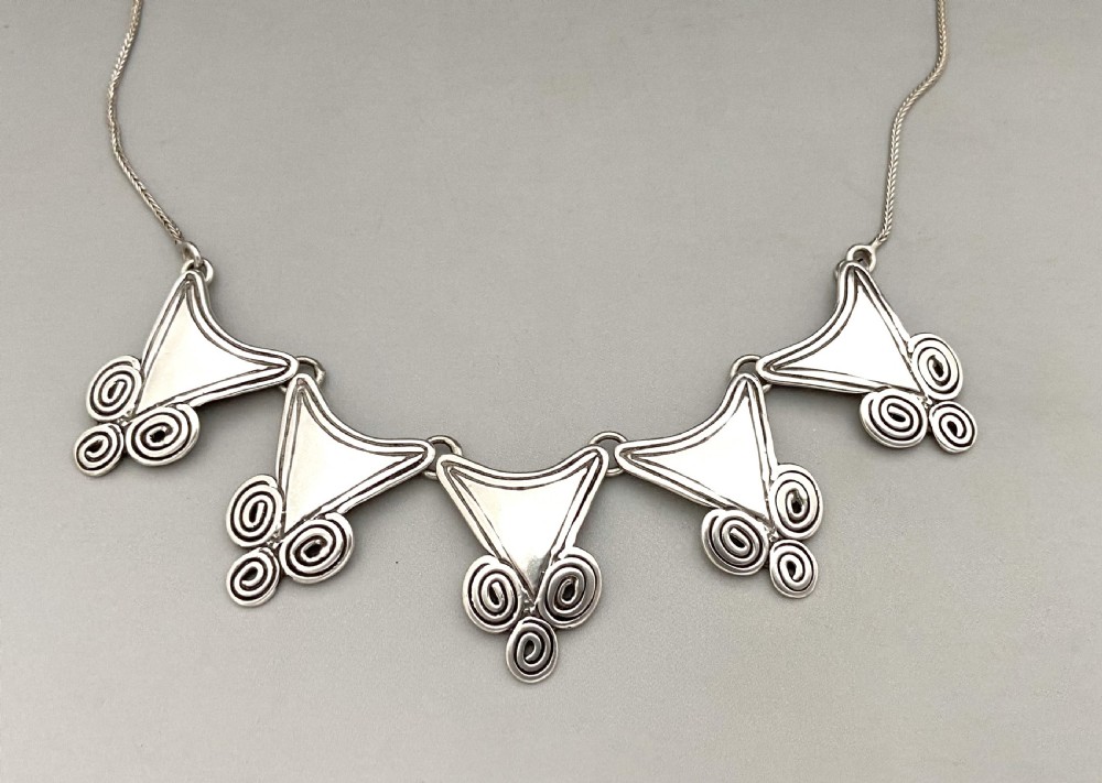 A mid-century silver necklace featuring five triangular linked panels with spiral scrollwork details suspended from a fine silver chain.