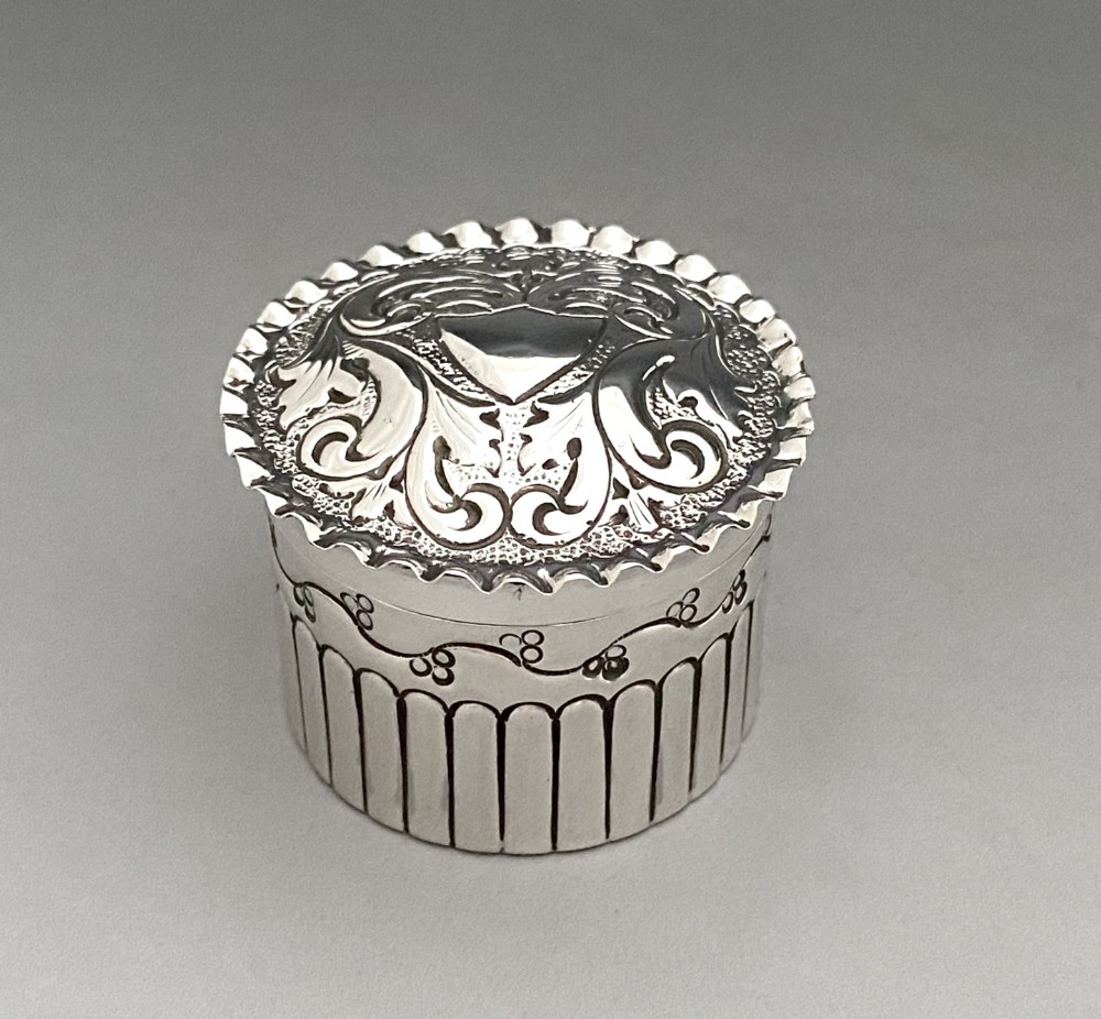 A circular Victorian sterling silver pill box decorated with foliate scroll engraving, an unengraved shield cartouche, a crimped rim, and vertical fluting around the base.