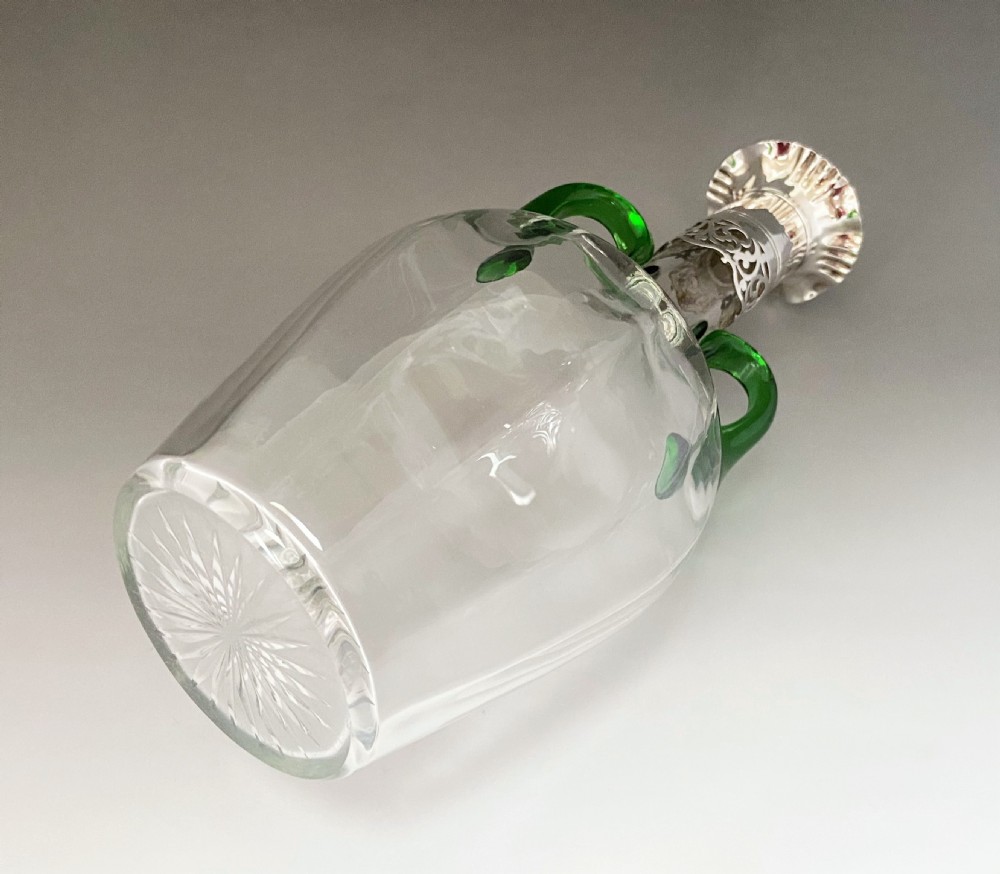 An Art Nouveau clear glass decanter featuring two applied green glass handles, a silver collar with openwork foliate motif, a scalloped silver rim, and a clear glass stopper. View V