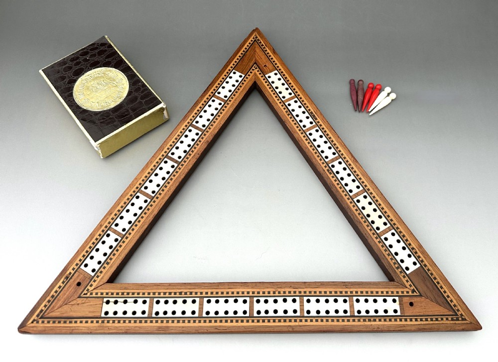 A mid-Victorian triangular wooden cribbage board with geometric inlaid borders, inset white scoring tracks, six coloured pegs, and a playing card box.