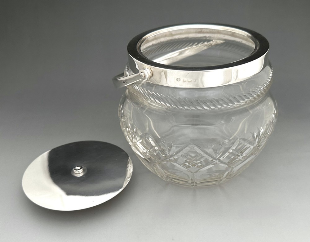 A George V clear cut glass biscuit barrel featuring a silver rim mount, a flat silver lid with central finial, and an arched silver swing handle. View VI