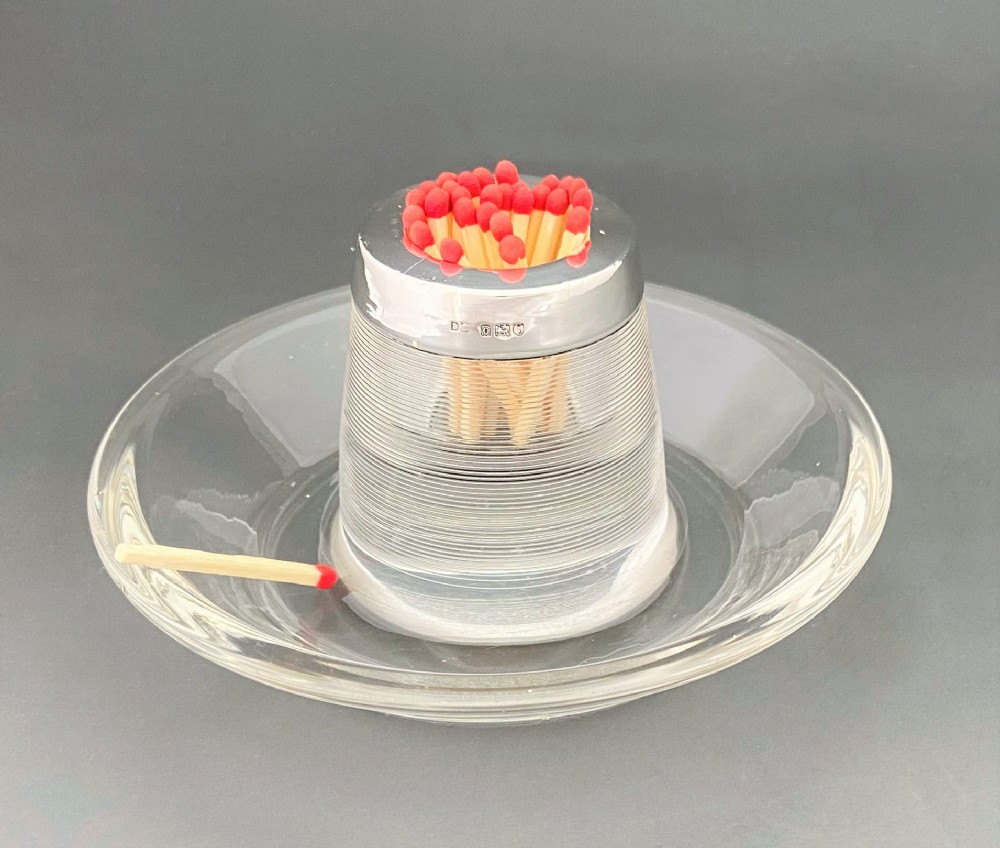 An Edwardian clear glass match striker featuring a ribbed cylindrical body, wide saucer base, and a hallmarked sterling silver collar holding red-tipped matches.