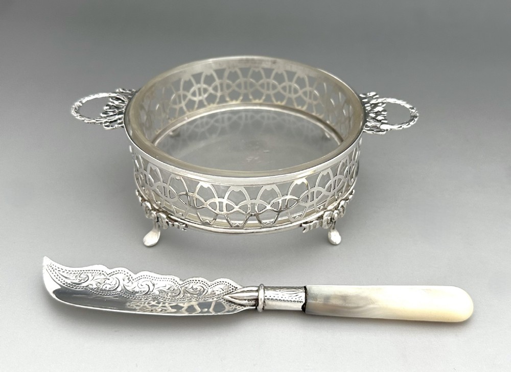 A circular pierced silver butter dish with glass liner and loop handles alongside an engraved silver butter knife with a mother-of-pearl handle. View II