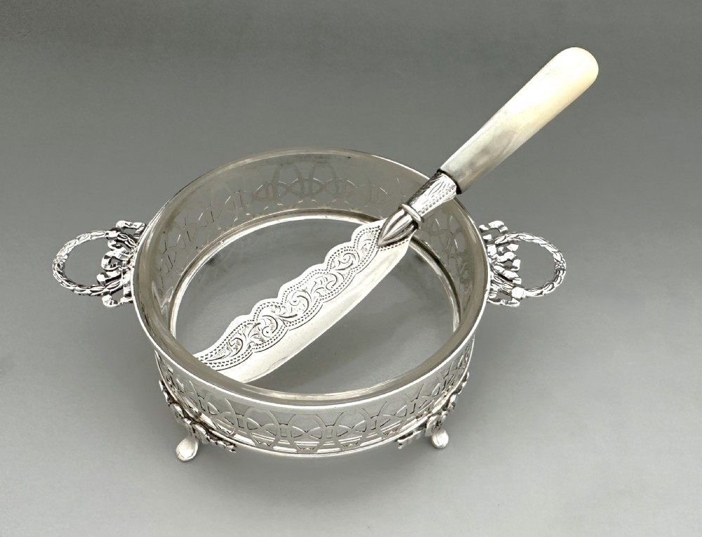 A circular pierced silver butter dish with glass liner and loop handles alongside an engraved silver butter knife with a mother-of-pearl handle. View III