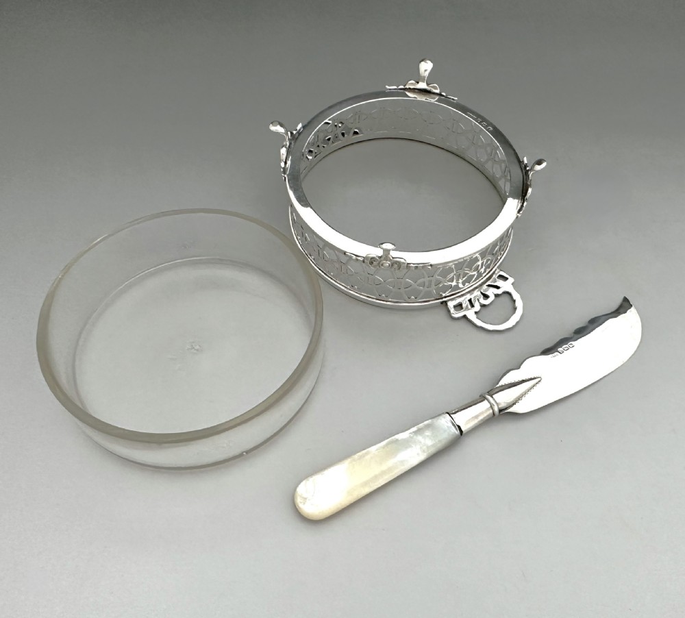 A circular pierced silver butter dish with glass liner and loop handles alongside an engraved silver butter knife with a mother-of-pearl handle. View VI