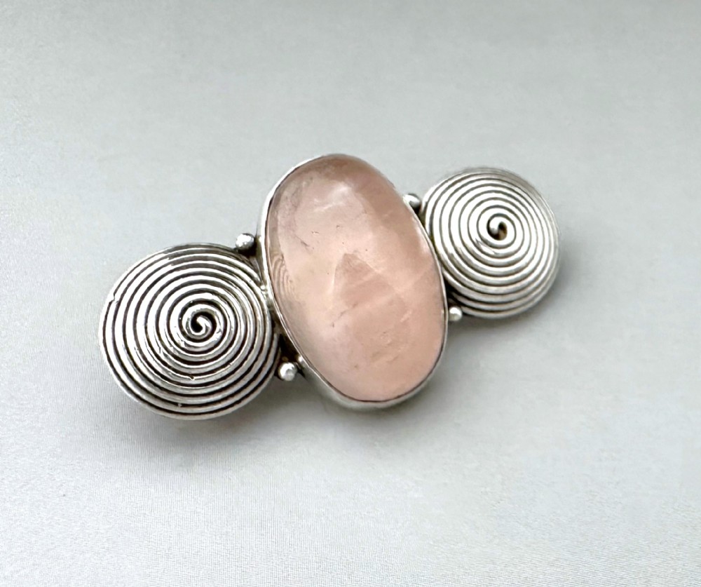 A silver brooch featuring a central oval cabochon rose quartz flanked by two spiral silver wire motifs.