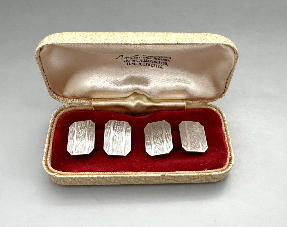 A pair of rectangular silver cufflinks with geometric engine-turned engraving displayed inside a cream fitted presentation box with red velvet lining.