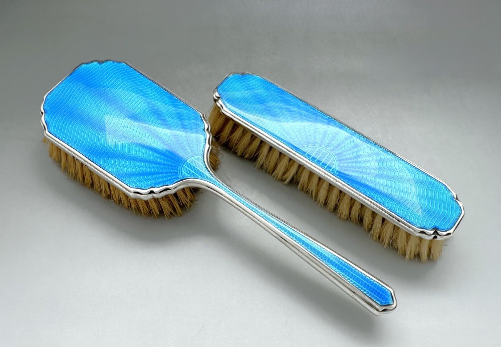 A pair of Art Deco silver vanity brushes featuring bright blue sunburst guilloche enamel panels and natural light bristles.