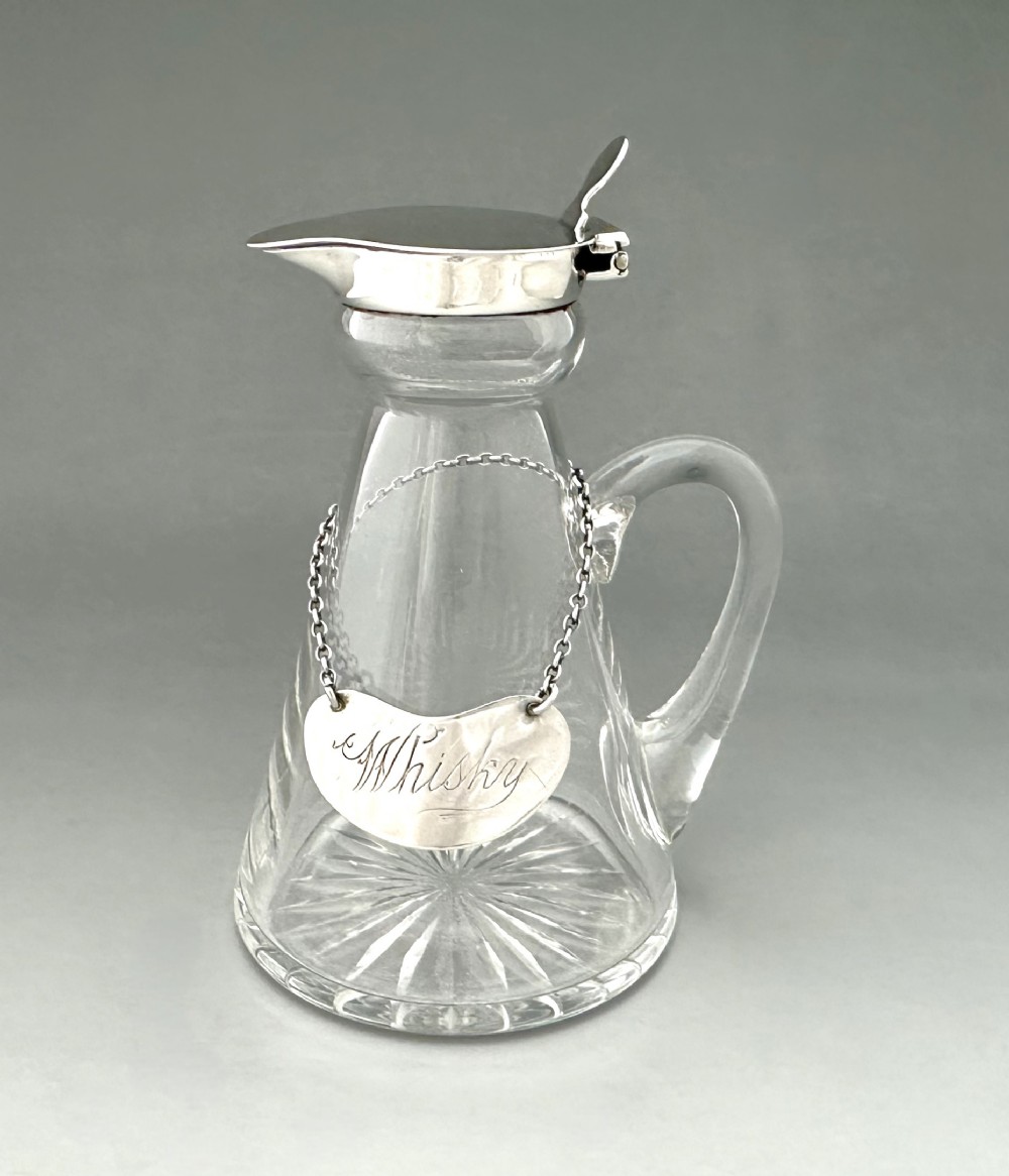A clear glass conical whisky tot with a star-cut base, a loop handle, a hinged sterling silver lid, and a silver label inscribed with Whisky.