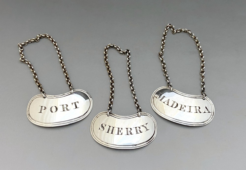 A set of three crescent shaped sterling silver decanter labels with chains, engraved with the words Port, Sherry, and Madeira.