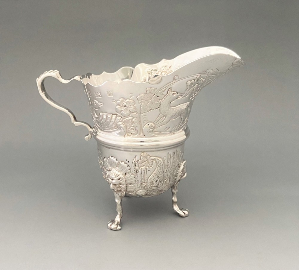 An antique sterling silver cream jug featuring chased animal and floral motifs, a scroll handle, and three lion mask feet.