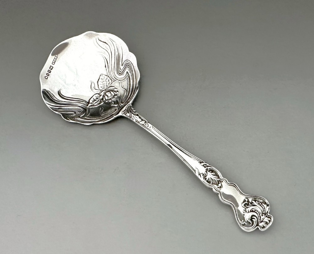 An antique Art Nouveau sterling silver preserve spoon featuring a scalloped bowl with an embossed water lily design and a hallmarked surface.