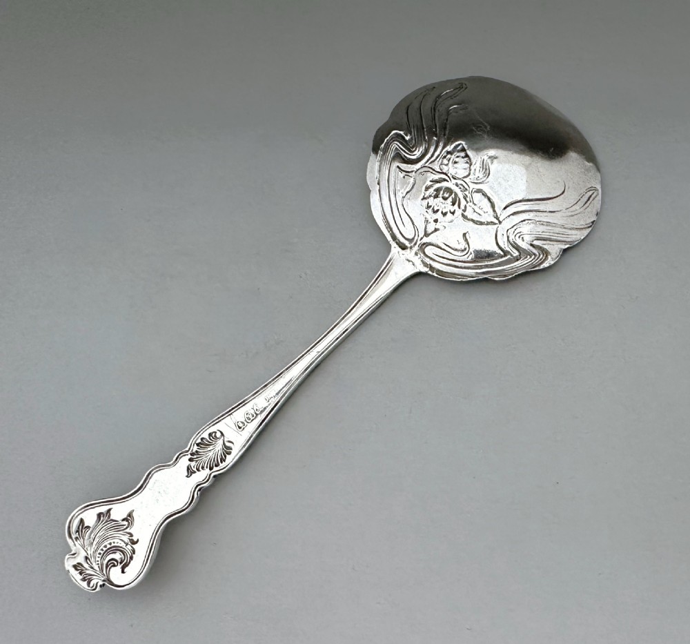 An antique Art Nouveau sterling silver preserve spoon featuring a scalloped bowl with an embossed water lily design and a hallmarked surface. View III