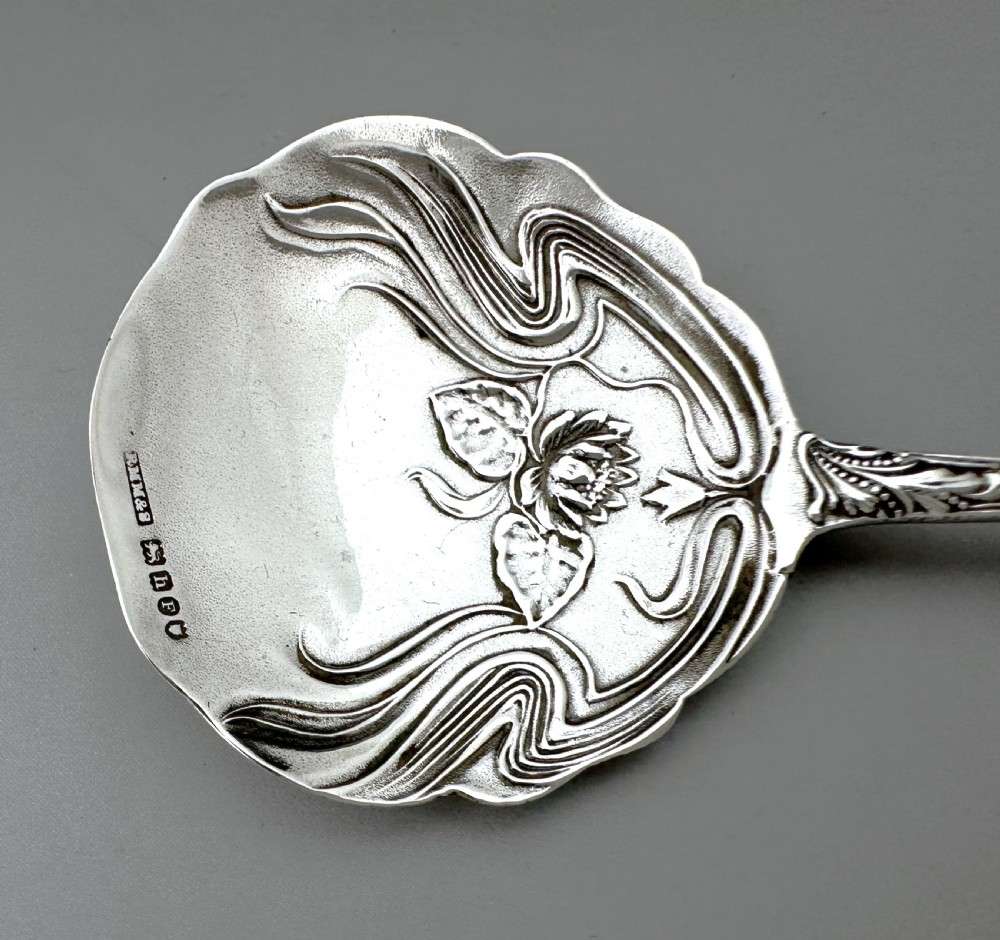 An antique Art Nouveau sterling silver preserve spoon featuring a scalloped bowl with an embossed water lily design and a hallmarked surface. View VI