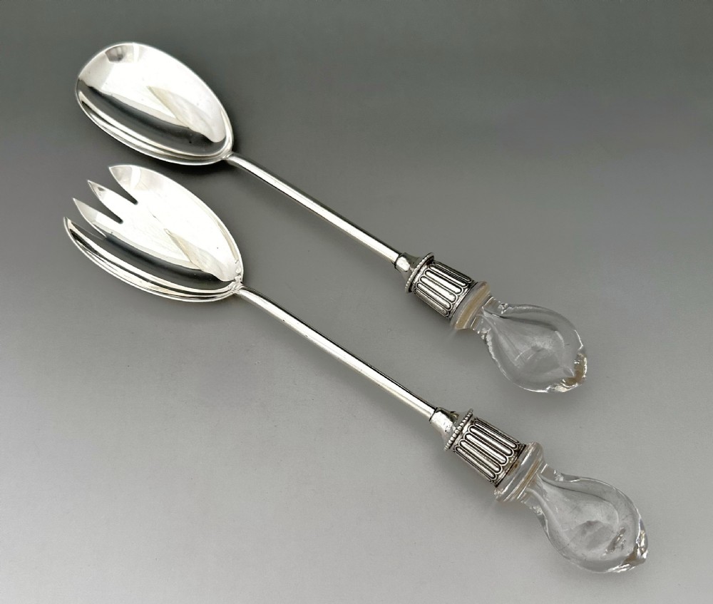 A pair of Edwardian silver salad servers featuring a rounded spoon and a three-pronged fork with decorative fluted ferrules and clear glass drop handles.