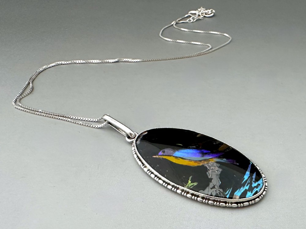 An oval Art Deco sterling silver pendant featuring an iridescent blue butterfly wing background with a reverse-painted bird motif, attached to a fine silver chain.