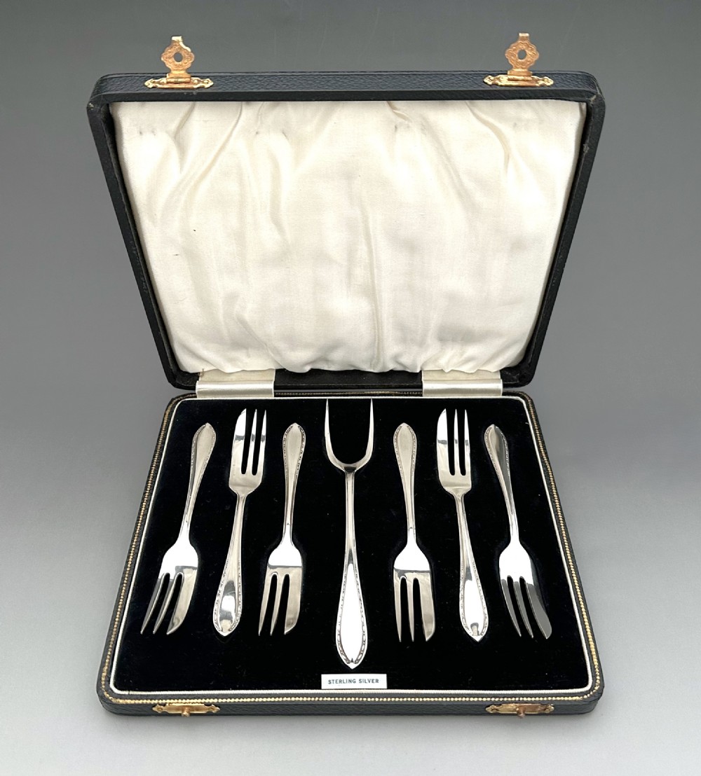 A cased set of six sterling silver cake forks and a central serving fork arranged in a black velvet-lined presentation box with a cream silk lid lining.