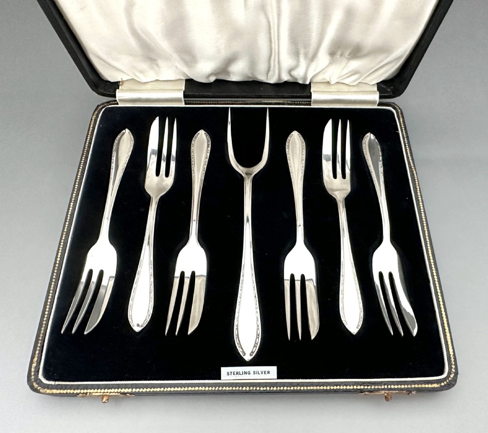 A cased set of six sterling silver cake forks and a central serving fork arranged in a black velvet-lined presentation box with a cream silk lid lining. View II