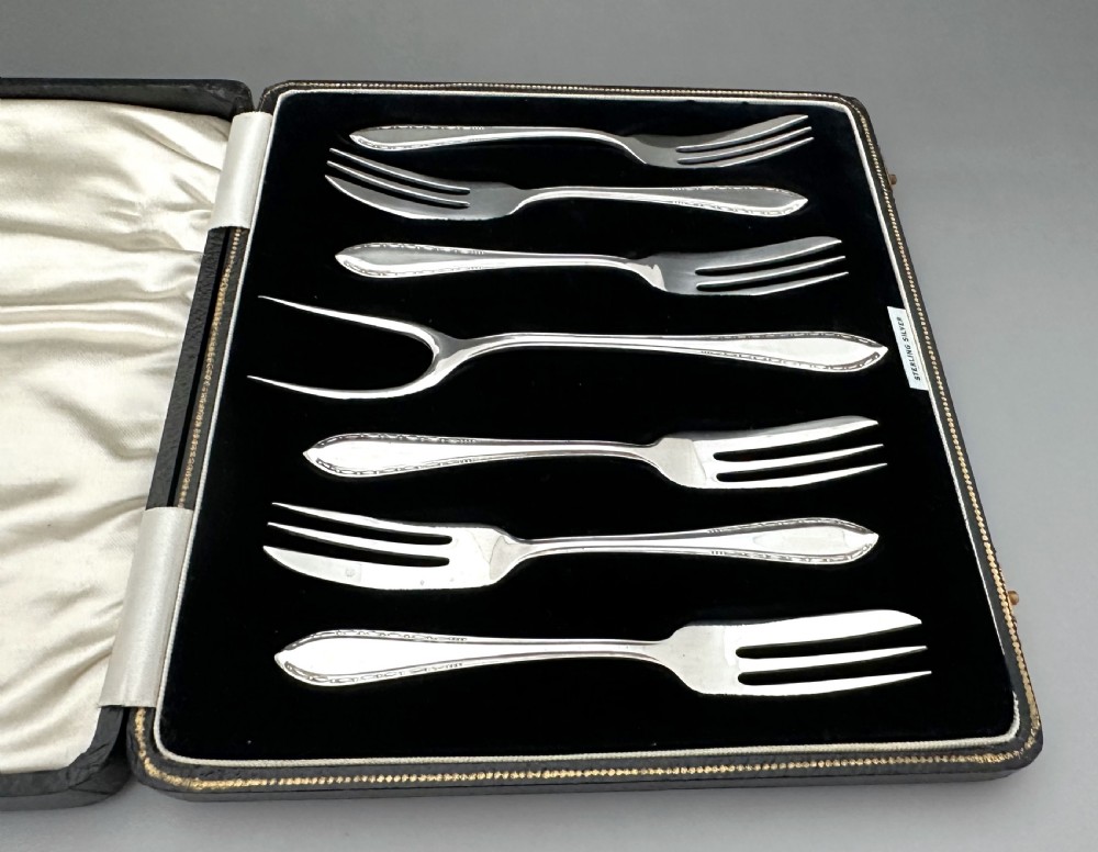 A cased set of six sterling silver cake forks and a central serving fork arranged in a black velvet-lined presentation box with a cream silk lid lining. View III