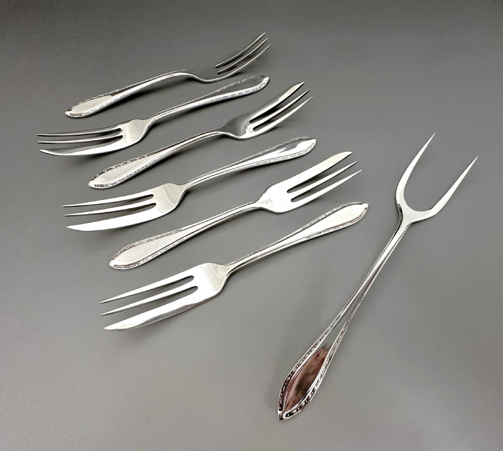 A cased set of six sterling silver cake forks and a central serving fork arranged in a black velvet-lined presentation box with a cream silk lid lining. View V