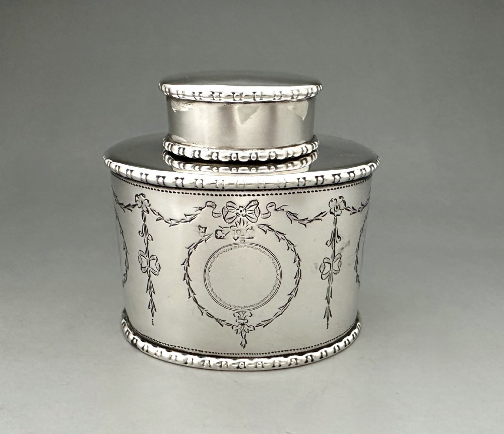 An oval sterling silver tea caddy featuring engraved floral garlands, ribbon swags, a vacant cartouche, and beaded borders.